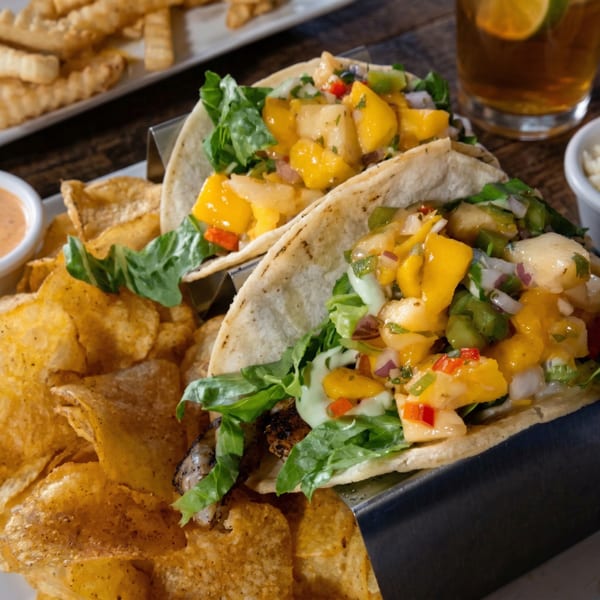 Two fish tacos with mango salsa and lettuce, served with potato chips and a drink.