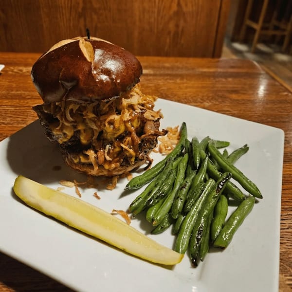 A pulled pork sandwich on a pretzel bun with crispy onions, green beans, and a pickle spear.